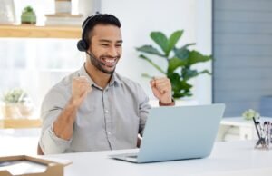 Een man met een headset zit aan een bureau, glimlacht en steekt opgewonden zijn vuisten omhoog terwijl hij naar een laptop kijkt.