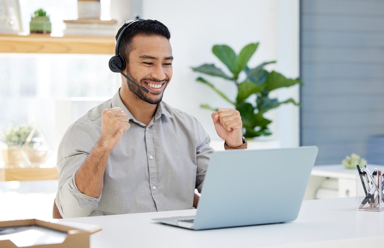 Een man met een headset zit aan een bureau, glimlacht en steekt opgewonden zijn vuisten omhoog terwijl hij naar een laptop kijkt.
