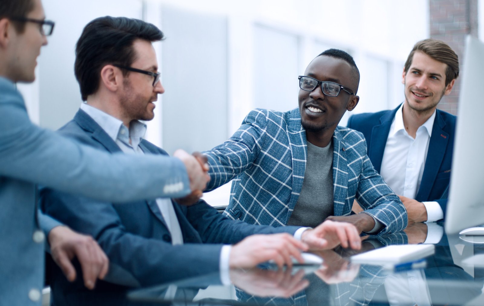 Vier mannen in zakelijke kleding zitten aan een glazen tafel. Twee van hen schudden elkaar de hand en glimlachen, wat duidt op een professionele ontmoeting of overeenkomst.
